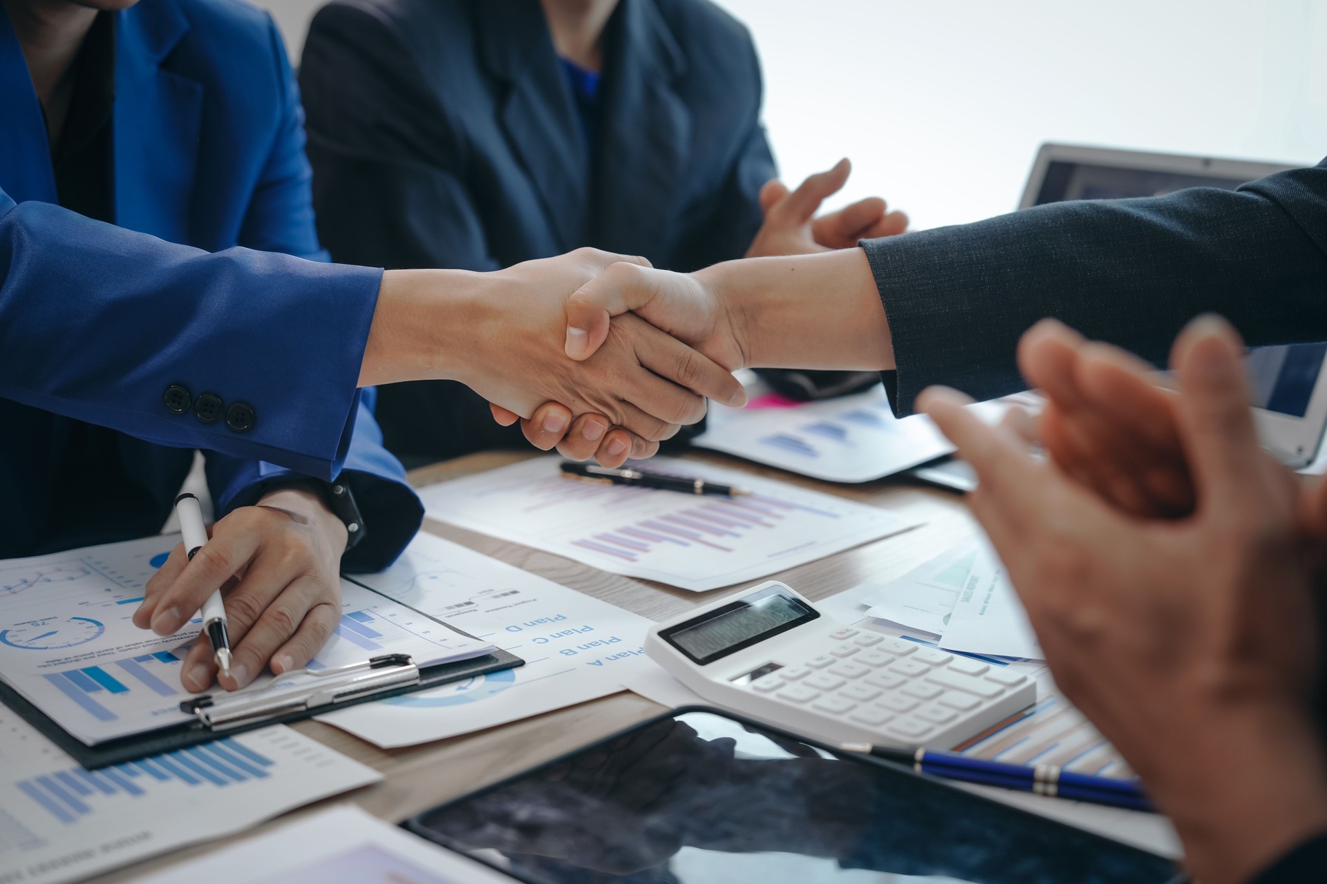 In a business team meeting, success is celebrated with a handshake. Hands close up, dressed in formal suits, working at desks with financial papers, calculators, and laptops.